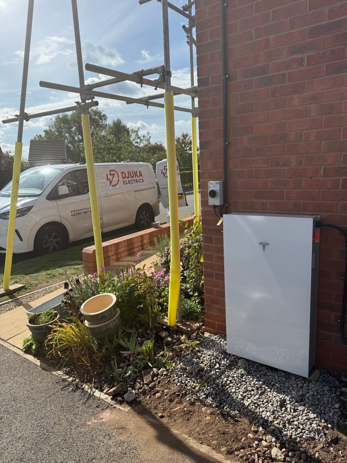 Tesla Powerwall installed on the side of a brick home with the Djuka van visible behind